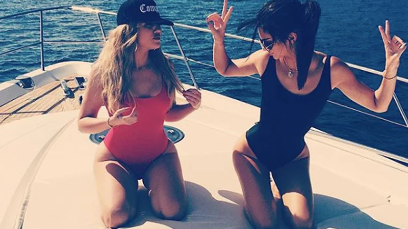 Two women in high-cut one-piece swimsuits kneeling on a yacht deck showing hip cleavage