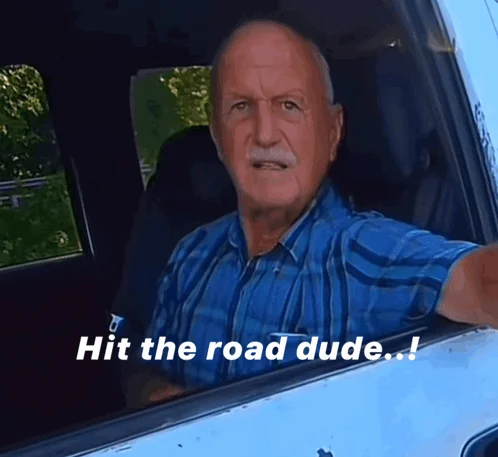 Older mustached man in a blue plaid shirt leaning out of a white pickup truck window, with the caption Hit the road dude.