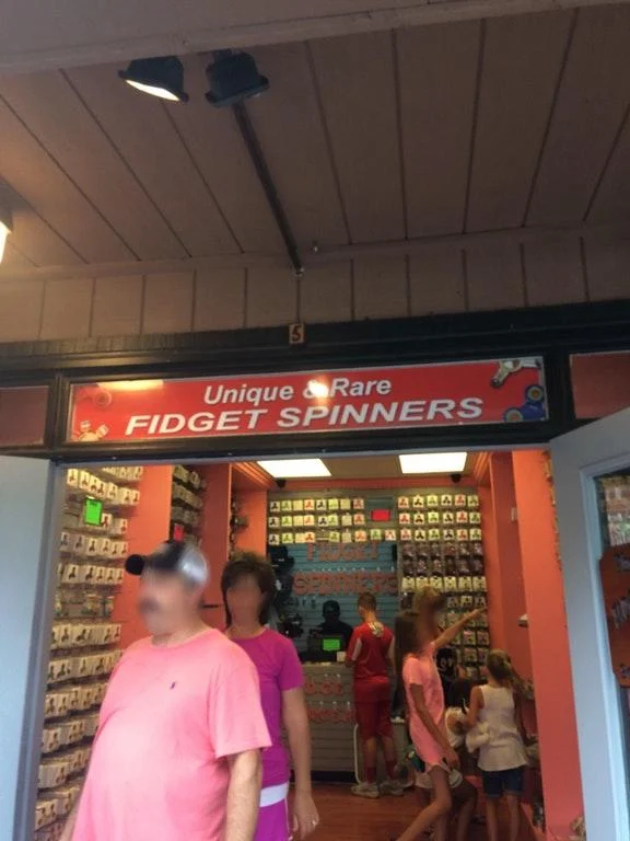 "How Do You Do, Fellow Kids?" — Storefront sign reading 'Unique and Rare Fidget Spinners' with shoppers inside