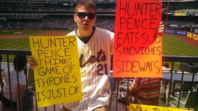 Hunter Pence Signs — Citi Field daytime photo of fan holding yellow sign about Game of Thrones and orange sign about eating sandwiches sideways.