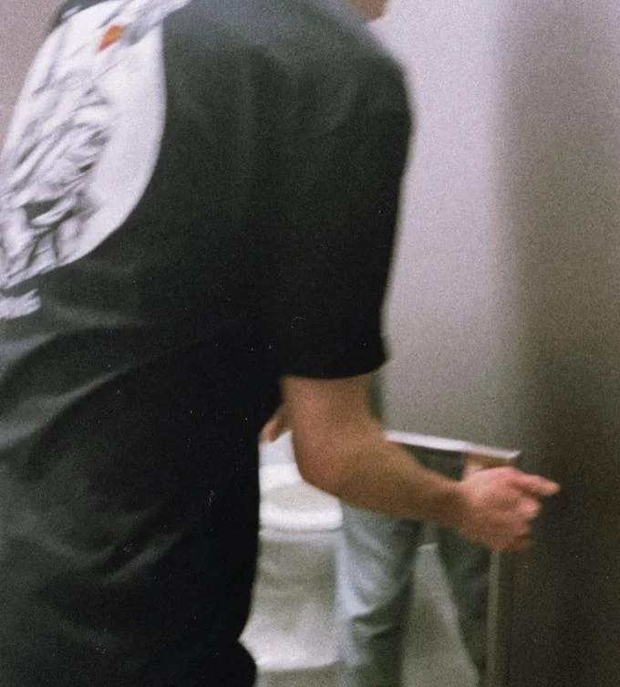 Hunter Schaffner and Holden Goyette Mirror Incident — Back view of a person in a bathroom stall holding a mirror fragment near a toilet