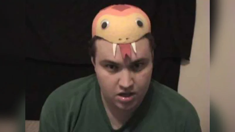 Im A Snake — Man in green shirt wearing white and pink snake hat with fangs, staring at camera with earnest expression against dark background.