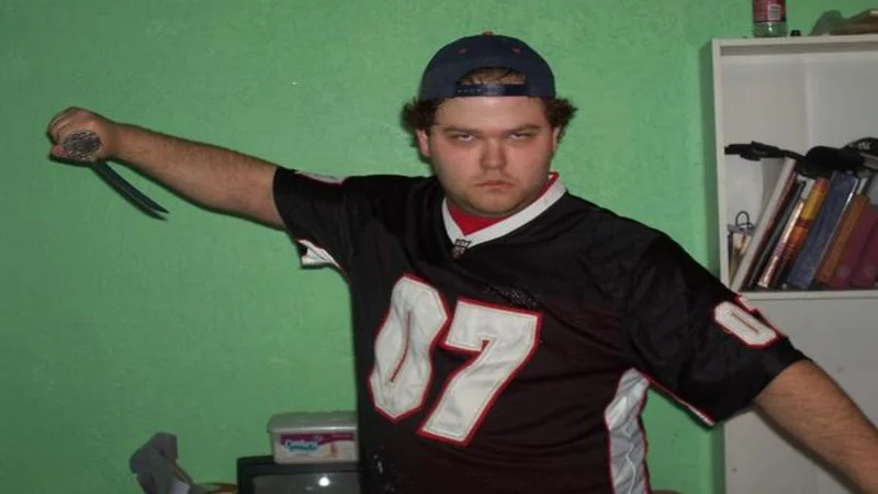 Internet Tough Guy — Heavyset man in sports jersey and backwards cap thrusting scissors toward camera in bedroom against green wall.