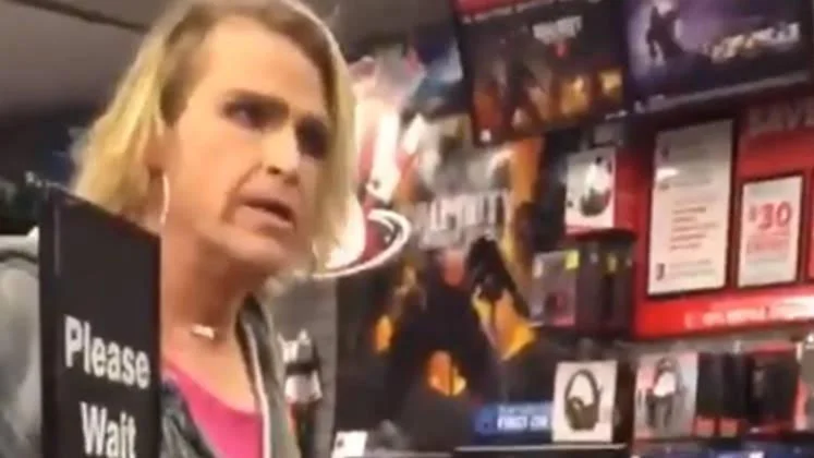 It's Ma'am — Screenshot of a person with long blonde hair standing at a GameStop counter next to a 'Please Wait' sign and video game displays.