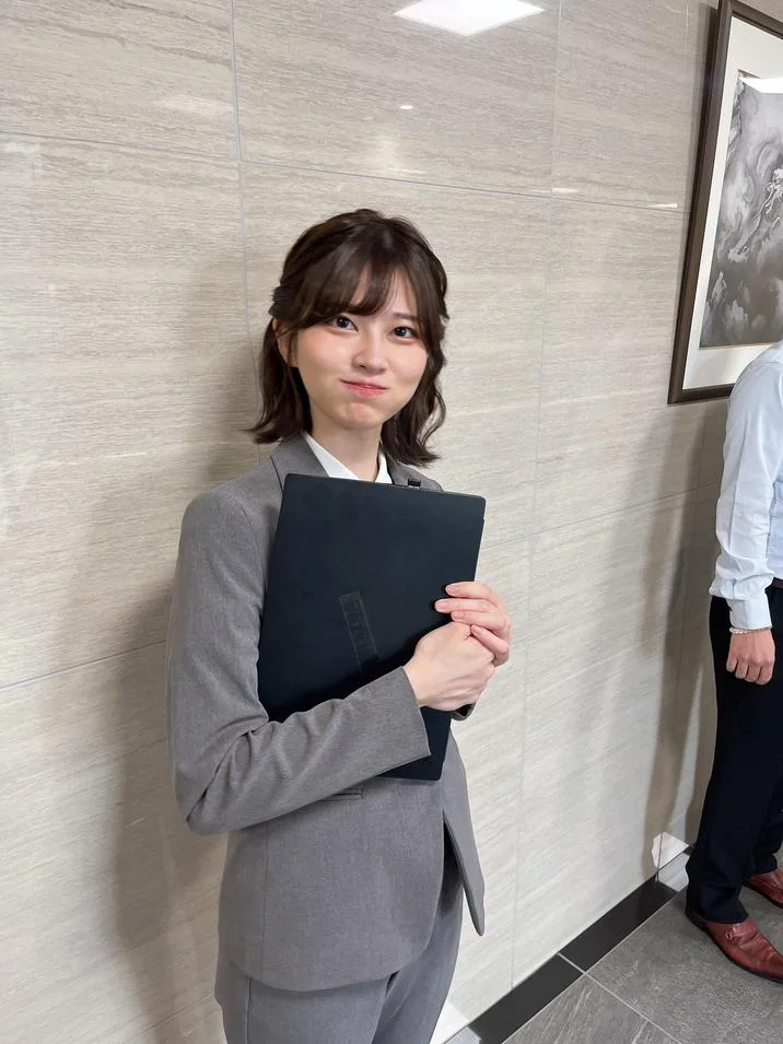 Japanese Salarywoman Saori Araki — A Japanese woman in a gray tailored business suit standing and holding a black laptop with a puffed-cheek smile.
