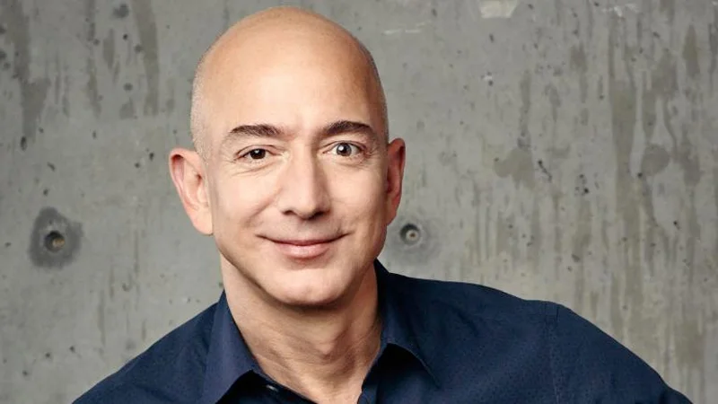 Portrait photo of Jeff Bezos smiling in a dark shirt against a concrete wall