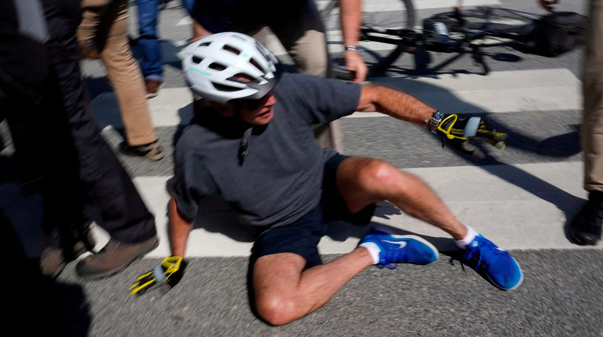 Joe Biden Falls Off Bike — Joe Biden sitting on pavement after falling off his bicycle while wearing a helmet, dark shorts, and blue sneakers.