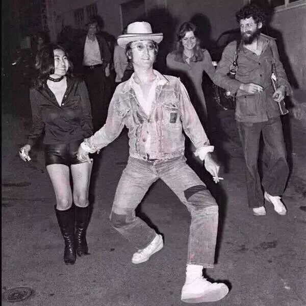 The Absolute Madman — Black and white photo of John Lennon doing a silly walk on a street with Yoko Ono beside him
