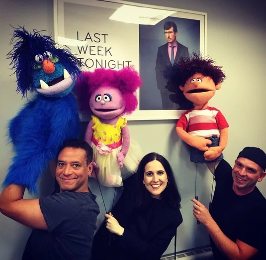 Puppeteers with colorful puppets posing in front of a John Oliver Last Week Tonight poster