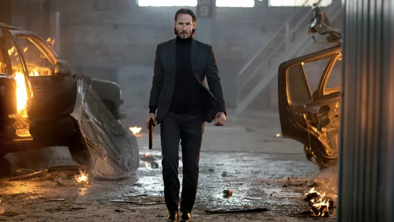 Keanu Reeves as John Wick in black suit walking with a pistol through a warehouse with cars on fire