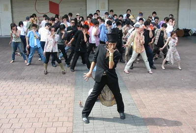 Jojos Pose — Dozens of fans in an outdoor plaza strike exaggerated angular JoJo's poses in unison with a figure in black suit and top hat leading.
