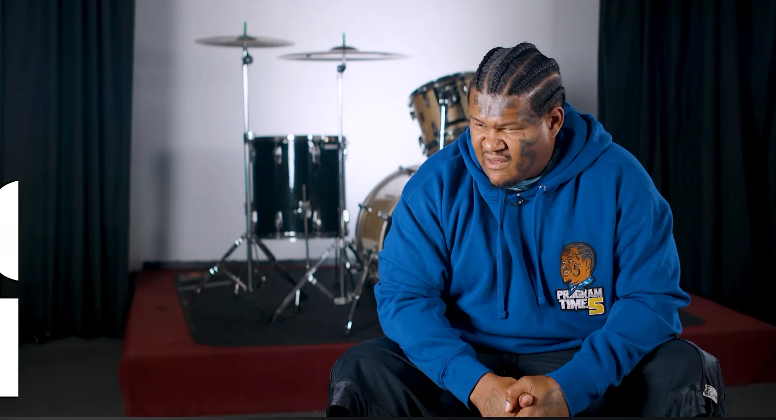 Kalani Bear, That's My Baby — Man in blue Program Time 5 hoodie with cornrow braids sitting in front of a drum kit during an interview