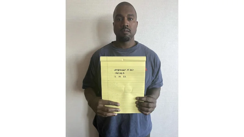 Kanye West Holding Notepad — Kanye West in navy blue t-shirt against white wall holding yellow legal pad with 'MY ACCOUNT IS NOT HACKED 2 13 22' handwritten.