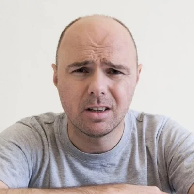 Karl Pilkington with a puzzled expression wearing a grey t-shirt