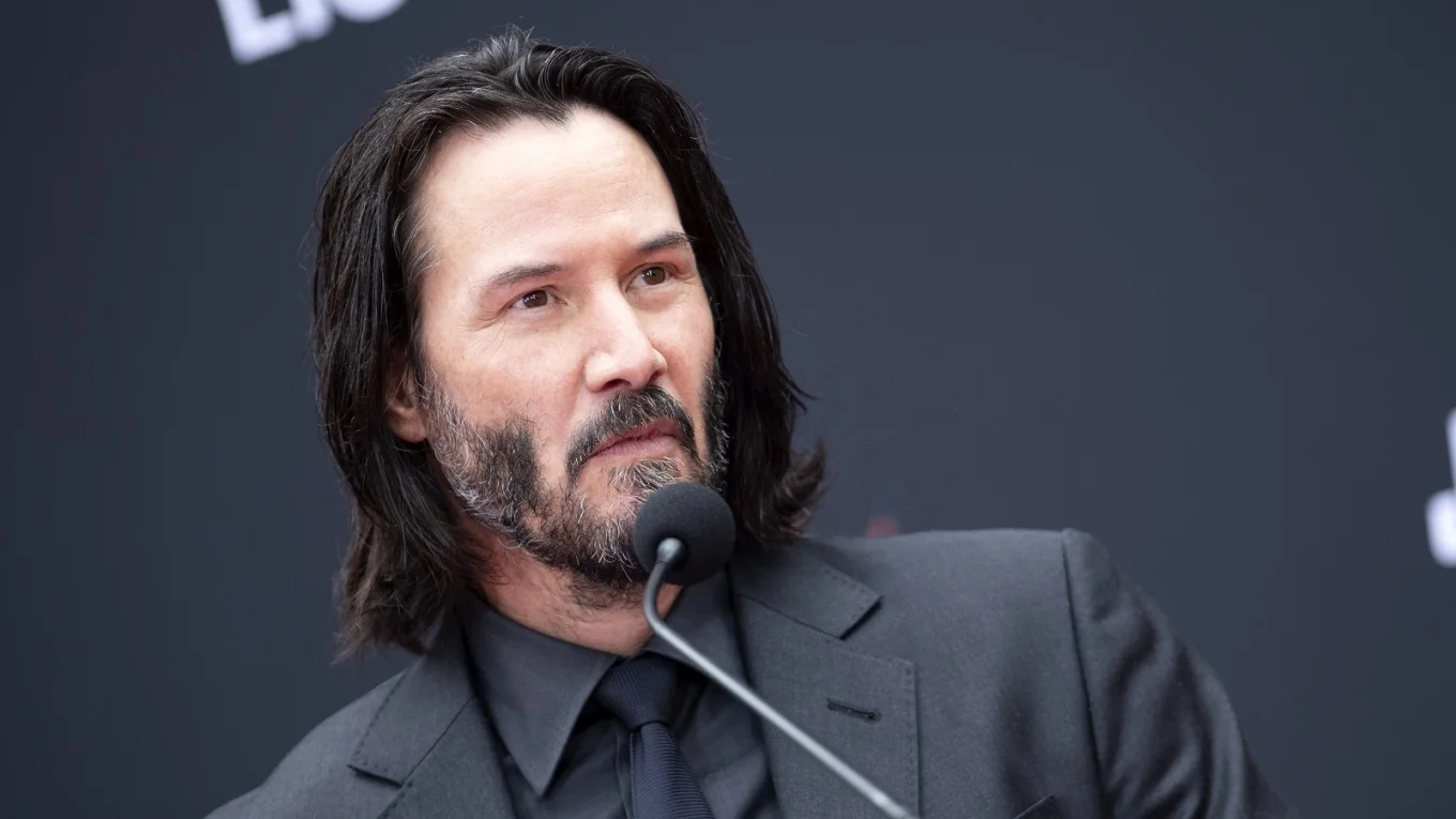 Keanu Reeves with long hair and beard speaking at a press conference microphone