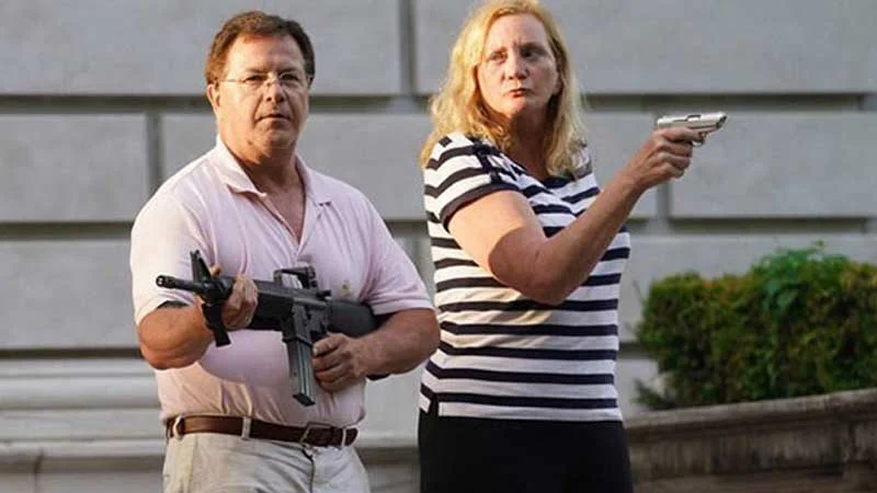 Ken And Karen St Louis Missouri Gun Couple — Middle-aged white couple standing outside a stone mansion, man in pink polo holding a rifle and woman in striped