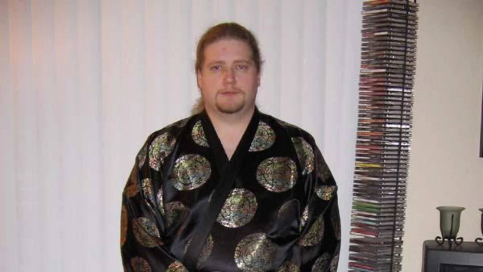 Ken-Sama — Man with ponytail and goatee wearing a black patterned kimono standing in a living room next to a CD tower.