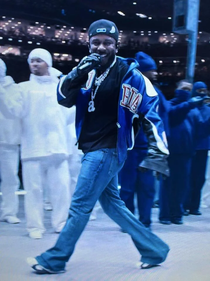 Kendrick Lamar Say Drake — Kendrick Lamar performing at Super Bowl LIX holding microphone, wearing blue SB-branded jacket and hat.