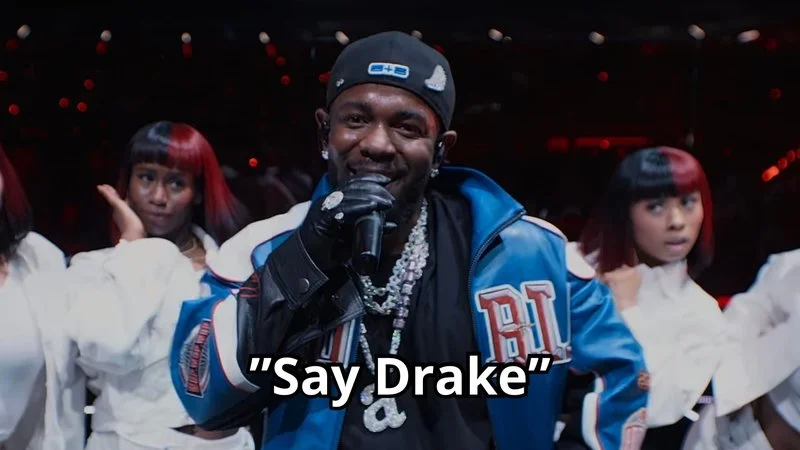 Kendrick Lamar Say Drake — Close-up of Kendrick Lamar performing at Super Bowl with prominent smile wearing blue SB jacket and chains.