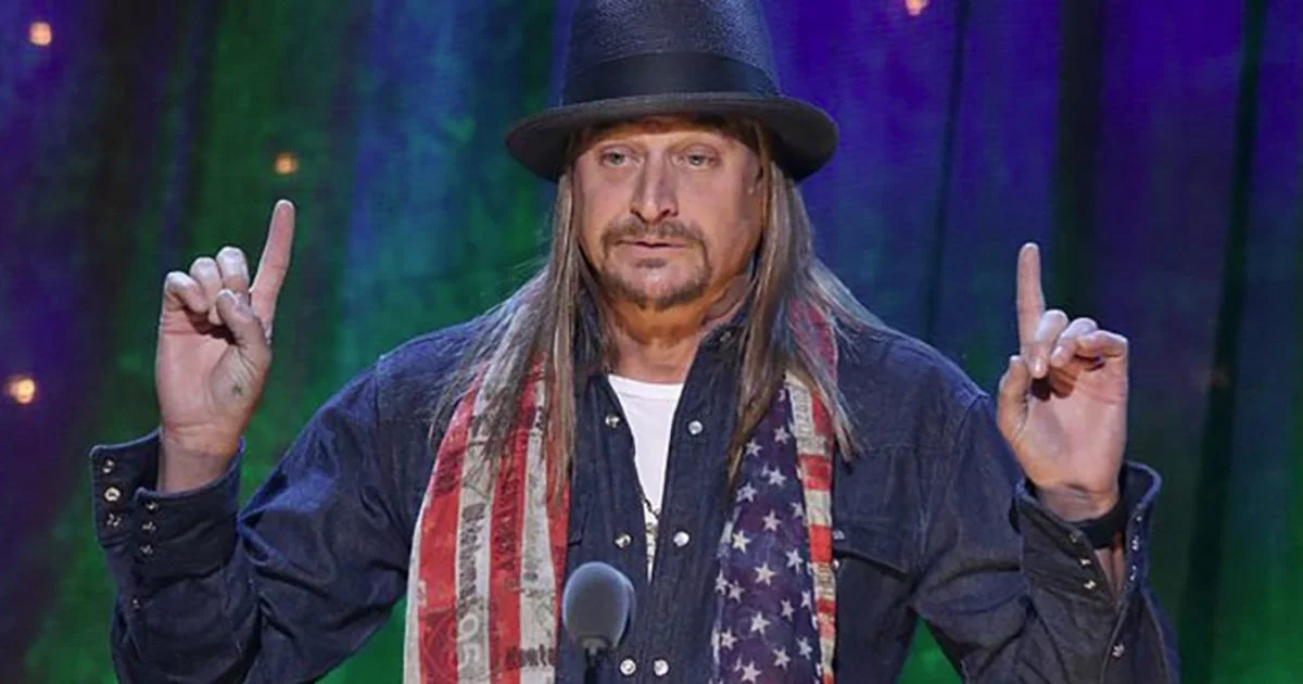 Kid Rock on stage in fedora and American flag scarf pointing both index fingers up