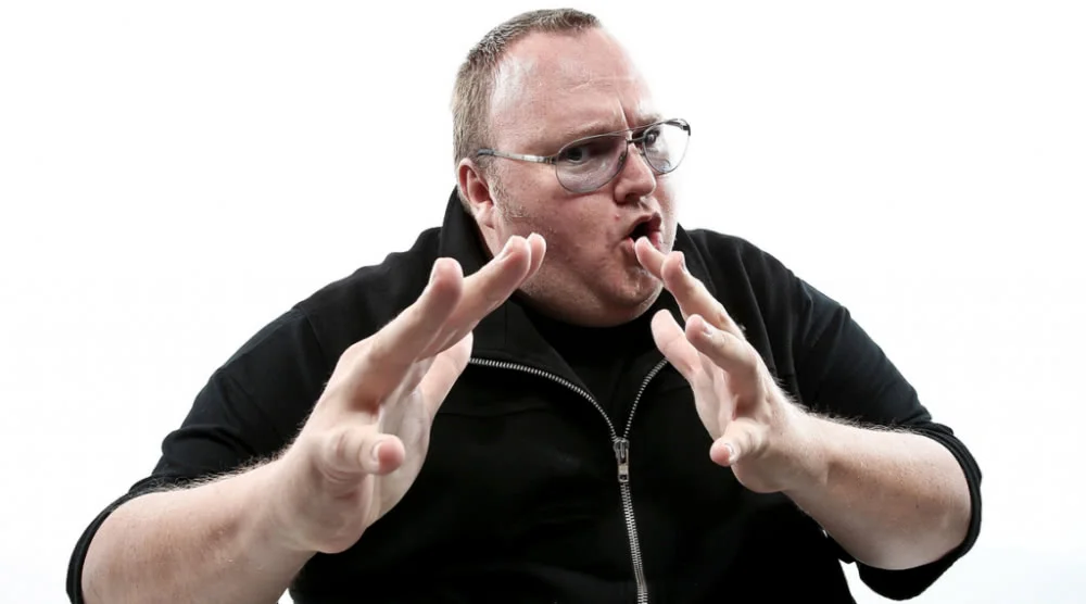 Kim Dotcom gesturing with both hands in a black jacket against a white background
