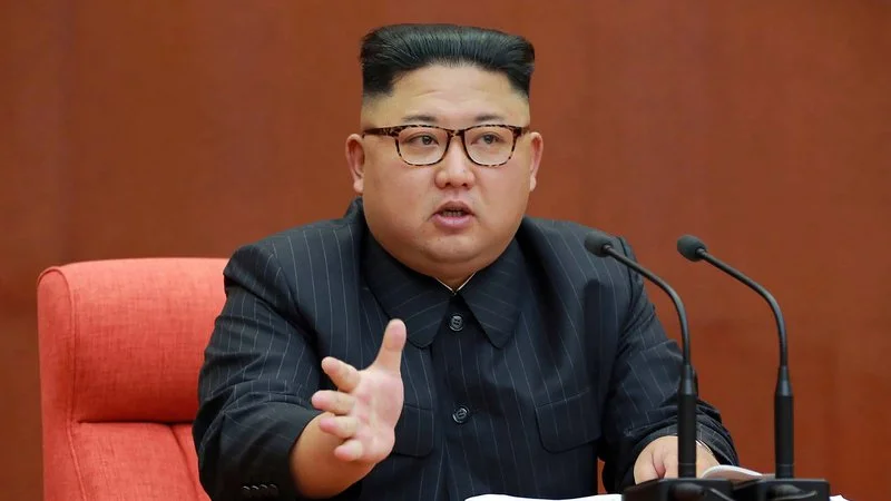 Kim Jong-un in glasses and black suit gesturing while speaking at a podium