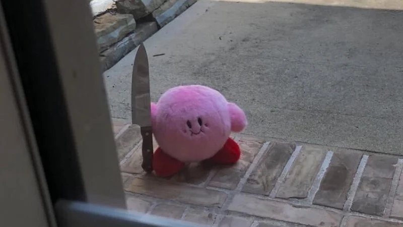 Kirby With A Knife — Pink Kirby plush toy holding a kitchen knife on a brick porch outside a door.