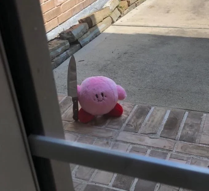 Kirby With A Knife — Uncaptioned photo of a Kirby plush holding a kitchen knife on a brick porch seen through a doorway.