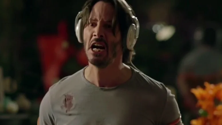 Knock Knock — Keanu Reeves in white headphones shouting angrily while wearing a dirty gray t-shirt