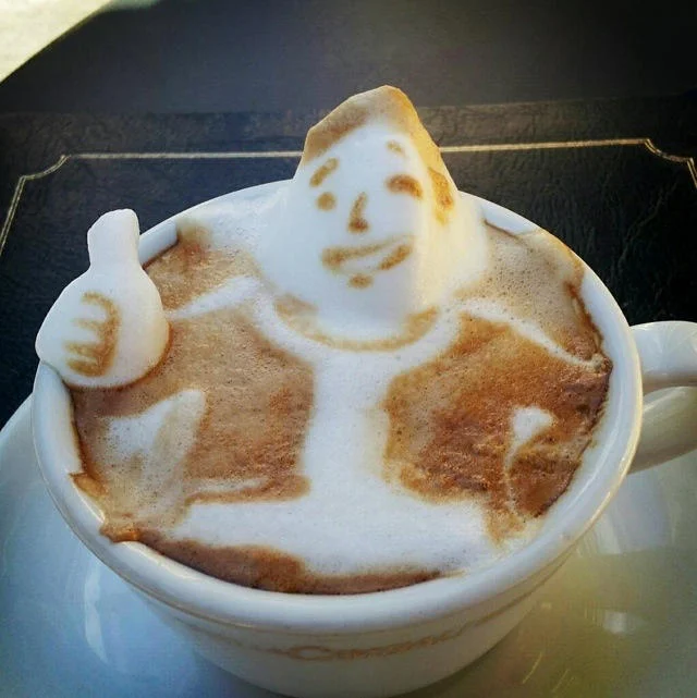 3D latte art of a smiling man giving a thumbs up rising from a coffee cup.