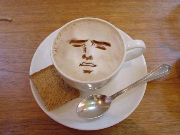Latte art of a stern man's face in a coffee cup with a spoon and biscotti on the saucer.