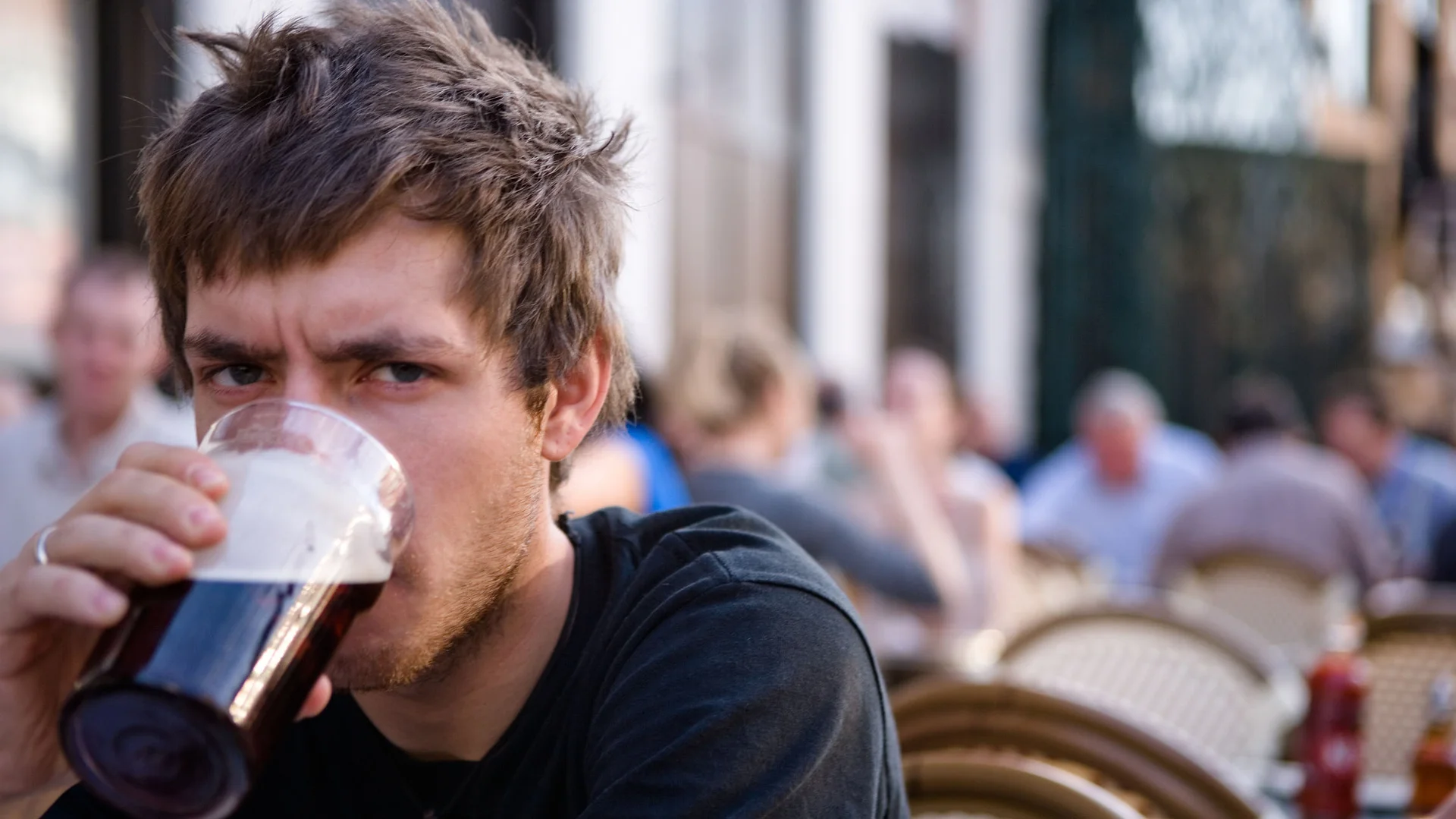Lazy College Senior — Young man with messy brown hair sipping a pint of dark beer at an outdoor cafe, staring blankly at the camera