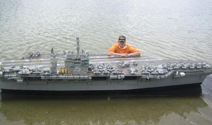Man standing next to a giant LEGO aircraft carrier floating in water