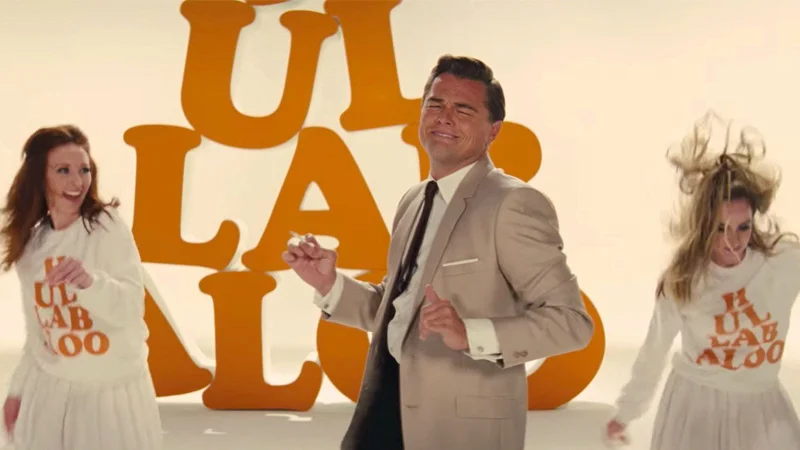 Leonardo DiCaprio dancing in tan suit between two women with orange 'Hullabaloo' letters in background