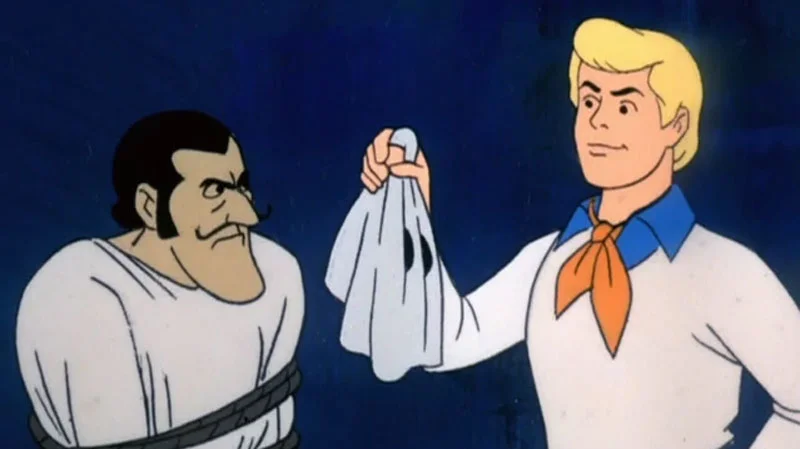 Let's See Who This Really Is — Fred from Scooby-Doo holding a white sheet mask next to an unmasked bearded villain