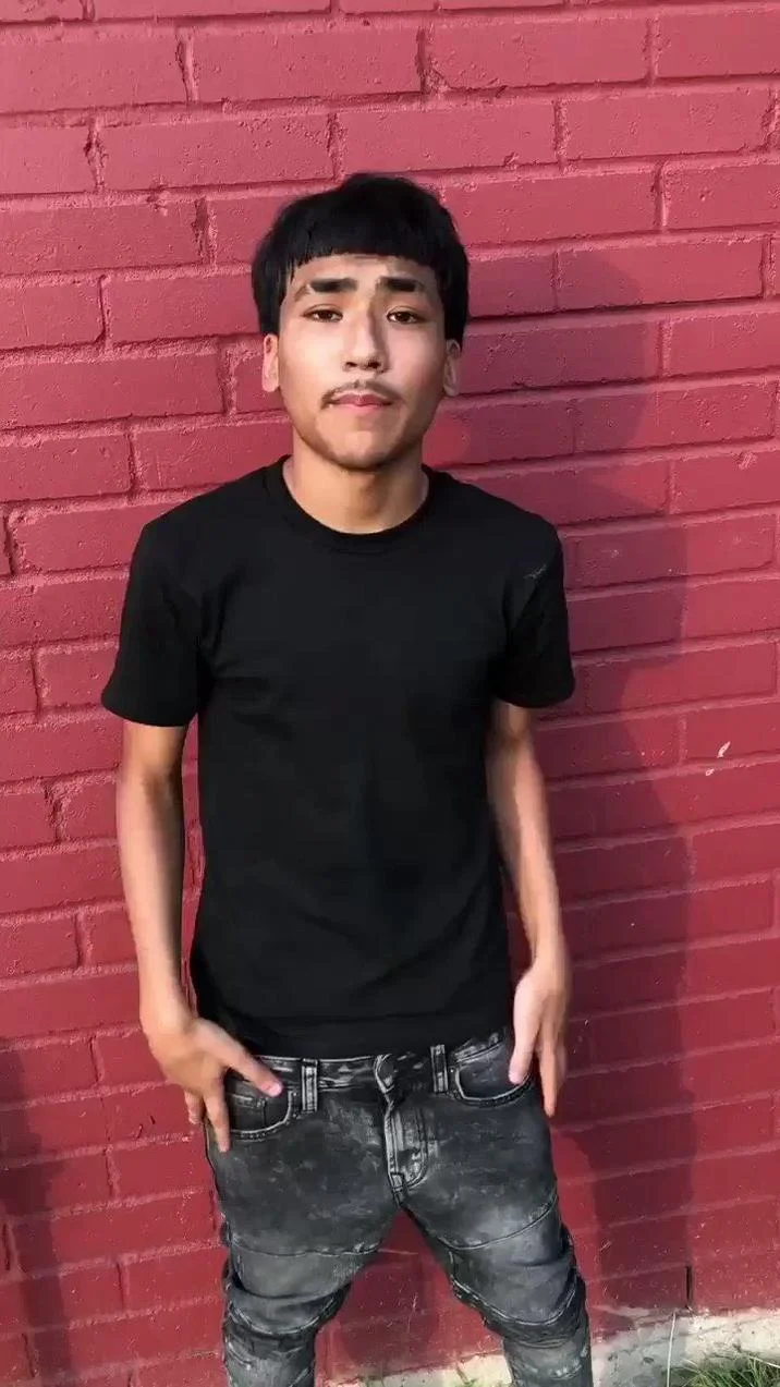 Lil G Photo Houston Resident — Lil G stands in a full-body pose against a red brick wall background, wearing a black t-shirt and distressed jeans with his