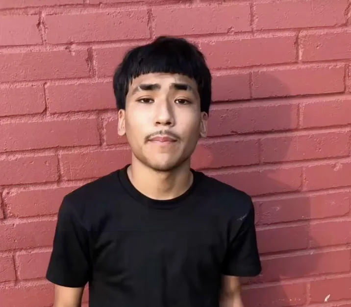 Lil G Photo Houston Resident — A clean headshot of Lil G featuring his distinctive Edgar haircut against the red brick wall background, wearing a black t-shirt.