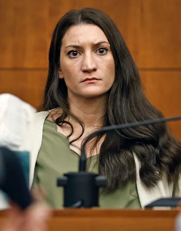 Lindsay Clancy Trial — Woman with long dark hair in a green top sits at a courtroom witness stand behind a microphone.