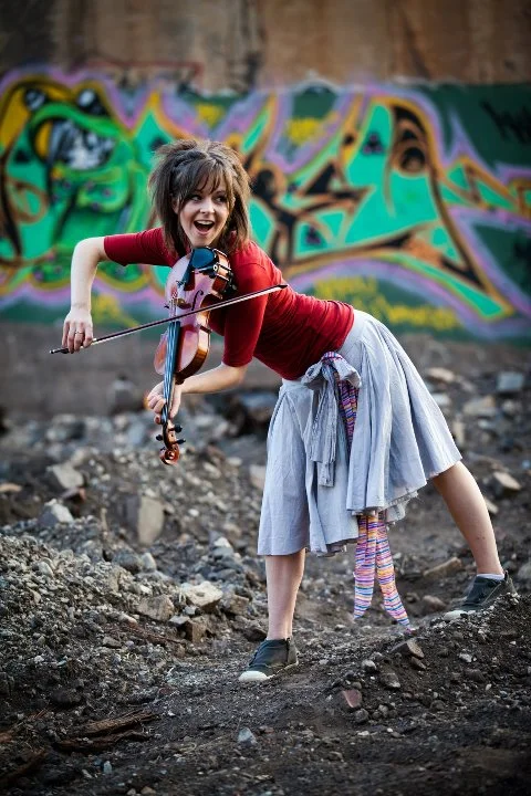 Lindsey Stirling dancing while playing violin in front of a graffiti mural