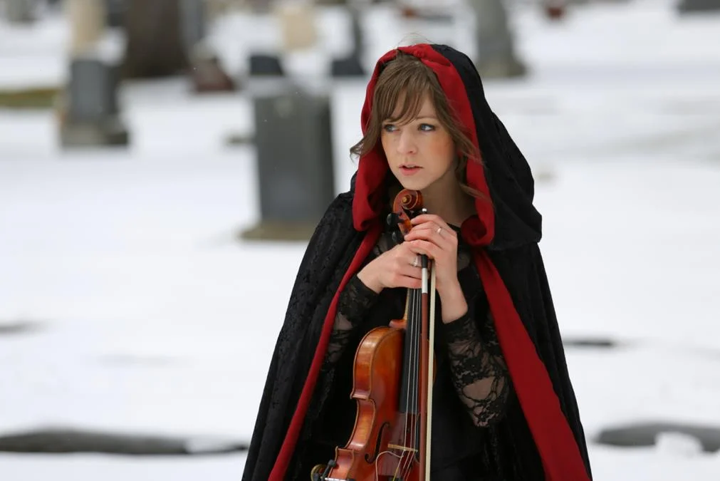 Lindsey Stirling in a red-lined black hooded cloak holding a violin in a snowy cemetery