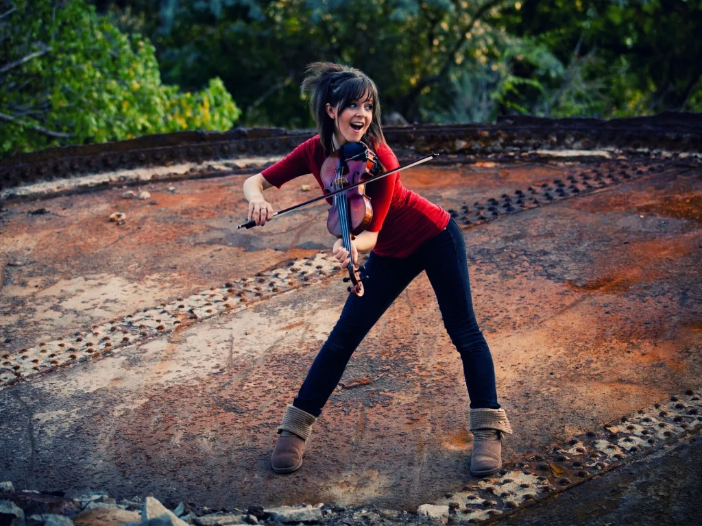 Lindsey Stirling playing violin in a red shirt on a rusty metal surface outdoors