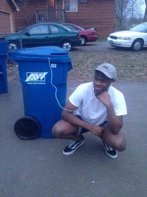 Listening to Trash — Young man crouched next to blue Allied Waste trash bin with earbud wires plugged into the can