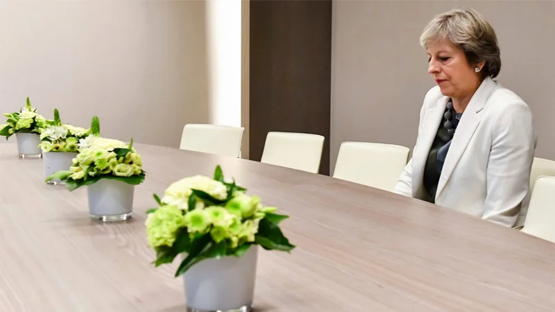 Lonely Theresa May — Theresa May sitting alone at the end of an empty conference table with flower centerpieces.
