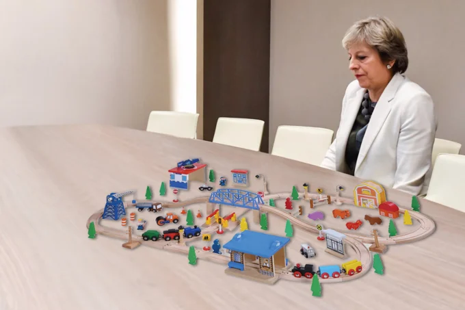 Lonely Theresa May — Theresa May seated at conference table with a wooden train toy set photoshopped onto the table in front of her.