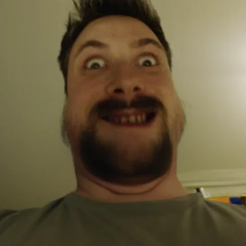 Luka Tim — Close-up selfie of a bearded man grinning widely with eyes open and teeth showing in a dim room.