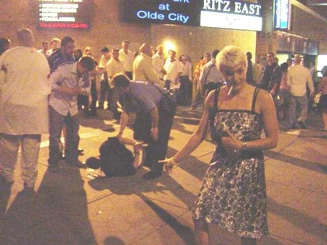 Lynndie England Pose — Man smoking a cigarette pointing and thumbs-upping at a woman collapsed on a sidewalk.