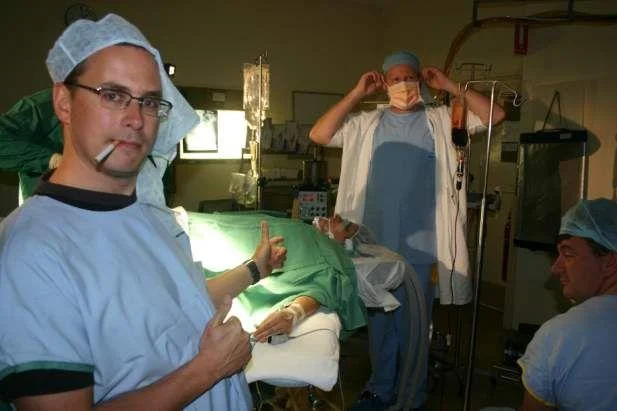 Lynndie England Pose — Doctor in scrubs with cigarette giving thumbs up and pointing at a patient in an operating room.