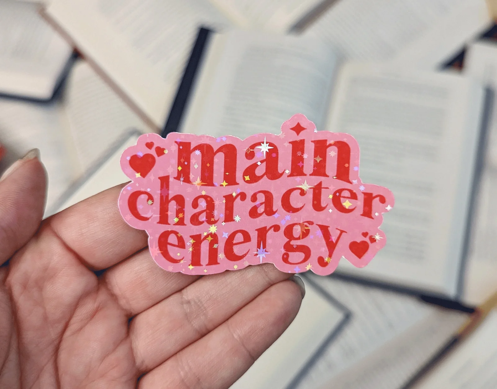 Pink 'main character energy' sticker with hearts and sparkles held over books
