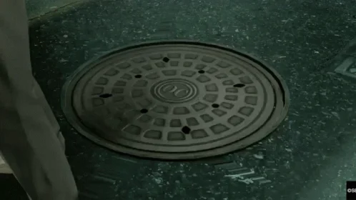 Majima Everywhere — A still frame from Yakuza Kiwami showing a closed manhole cover on a rain-slicked street, the tense moment before ambush.
