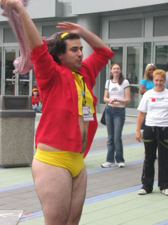 Man-Faye cosplayer in yellow crop top and shorts with red sash posing outdoors